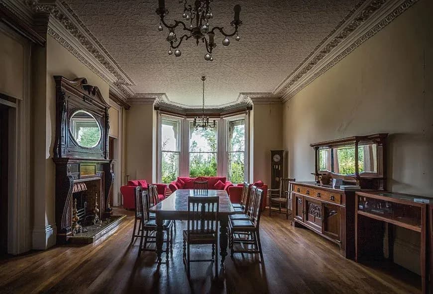 Elegant historic dining room with ornate fireplace, chandelier lighting, bay windows, red lounge seating and hardwood floors in a heritage home.
