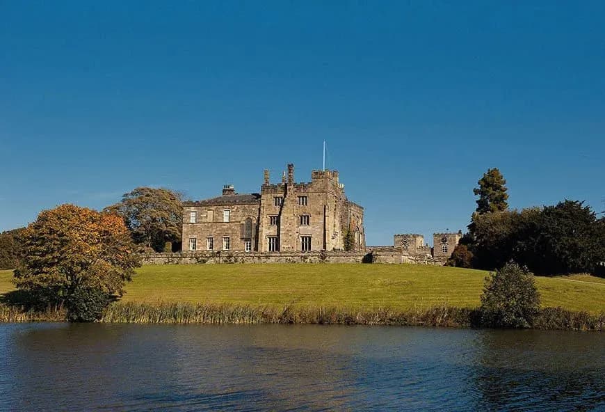 Large stone House on a hill with a lake in front of it.