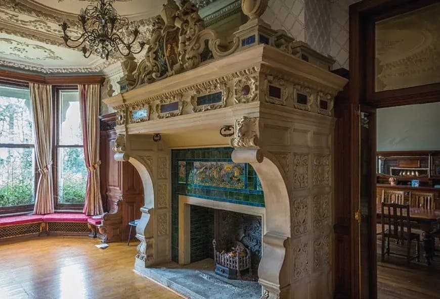Ornate historic living room with large carved stone fireplace, decorative tile surround, chandelier, bay windows and hardwood floors in a luxury heritage home.